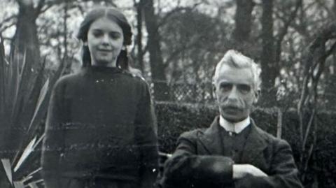 A black and white image of Elsa and William Hall. She is long dark hair pulled back from her face and is wearing a dark jumper over a dark skirt. He is sitting to her left and has white hair and a white moustache. He has a high white collar under a dark jacket and waistcoat and has his arms folded. Behind them is a garden.