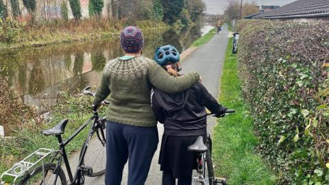 A mother stands with her back to the camera with her arm around her daughter's shoulders. They are standing next to the Union Canal and both are holding their bikes. The mum is on the left and is wearing a green knitted jumper and a colourful wooly hat and the daughter is wearing a cycling helmet and dark skirt and top.