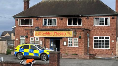 Shot of a fire-damaged building with a police car parked at the front. It has Georgian-style windows and a sign which says Golden Phoenix above the door.