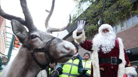 A reindeer is standing next to a waving Santa Claus during Middlesbrough Christmas parade. They are surrounded by security workers wearing high visibility vests. Several children are watching the event. 