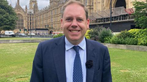 A smiling Charlie Dewhirst dressed in a blue suit standing outside the Houses of Parliament