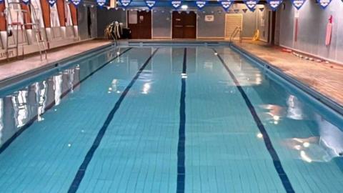 Interior of Ulverston Pool. It is a leisure pool with lanes marked with darker blue tiles at the bottom. There are flags hanging over the pool.