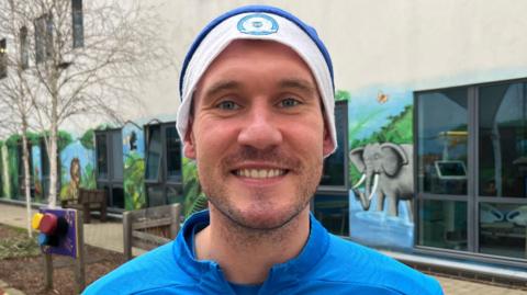 Jed Steer wearing a light blue tracksuit and matching blue Posh branded santa hat. He is smiling at the camera and stands outside the city hospital children's ward. He has blue eyes and stubble on his face. 