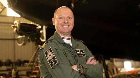 Squadron Leader Paul Wise stood wearing military uniform with his arms crossed in a warehouse in front of a military aeroplane, which is blurred in the background. He is bald and is smiling at the camera. 