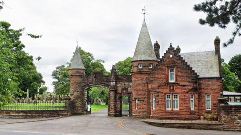 The gates of a cemetery with a red sandstone building adjoining an archway with turrets. 
