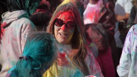 A woman covered in red, yellow and blue paint is smiling at the camera. She has long straight brown hair, sunglasses and is wearing a white t-shirt.