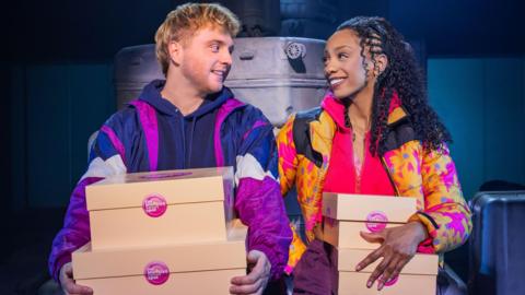Sam Tutty and Christiani Pitts, both holding cake boxes, in a theatre production. Sam has on a purple tracksuit and Christiani colourful tops and long hair. They are both looking at each other and smiling. 