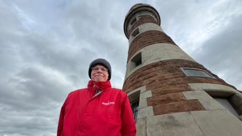 Stuart Robson stands in front of Roker Lighthouse. He is wearing a red jacket and black woolly hat.