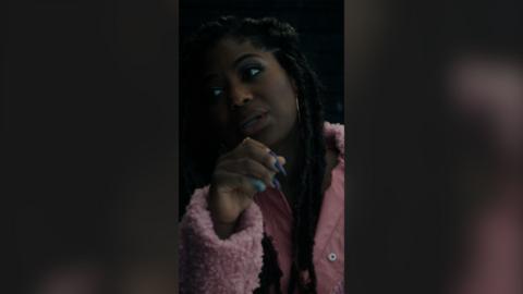 Sharla is speaking, her hand is partly in front of her face, she is wearing a pink coat