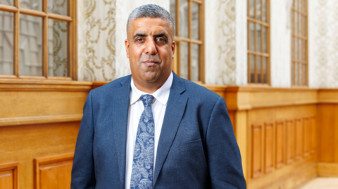 Professional portrait of a person wearing a blue suit, white shirt, and patterned blue tie, standing indoors in front of wood-paneled walls and large framed windows. The image is well lit, with the subject centered and facing the camera in a formal office or civic building setting.