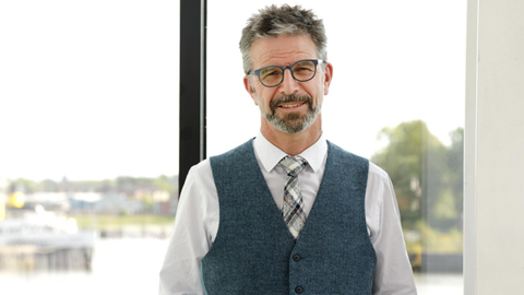 Prof Gordon Mackay - a man with glasses, a beard, moustache and dark/grey hair - standing in front of an office window, wearing a blue/grey waistcoat with a white shirt and patterned tie.