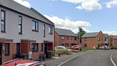 Modern semi-detached houses built since the 1980s along Bixton Close in Worcester. There are cars parked outside some of the buildings, and some of the home have lawns and flower pots outside.