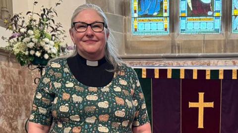 Rev Jane Lee with long grey hair wearing black glasses and a black and white clerical collar with a green cardigan over the top with patterns of sheep and flowers on it. She is standing inside St Wilfrid's Church in Melling near the altar.