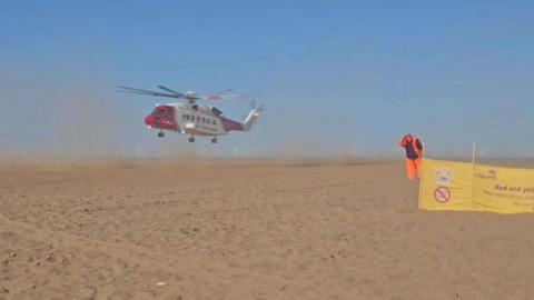 Sand is whipped up as a large red and white helicopter comes into land on a sandy beach under a blue sky. To the right, a man wearing orange hi-vis overalls bows his head as he stands next to a yellow Lifeguards sign.