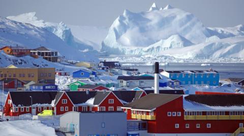 Houses and buildings sit in a snowy landscape with huge snow-covered mountains in the background.