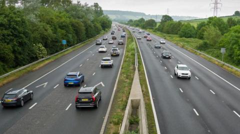 The M5 motorway near Weston-super-Mare, taken from above and showing all six lanes of traffic. There are several cars travelling in both directions and the motorway has trees on either side. Fields can be seen in the distance. 