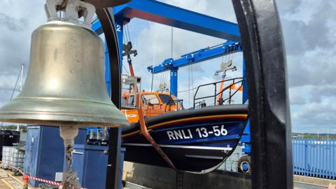 A large silver bell is in the top left-hand corner and behind it is an orange and blue RNLI lifeboat. It says RNLI 13-56 on it. It is suspended above the water on a crane. The sea can be seen in the distance.