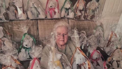 An elderly woman with short grey hair, glasses and a beige top. She is surrounded by teddy bears, grey in colour and all wrapped in polythene bags.