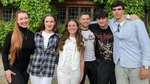 A mixed group of young men and women stand smiling in front of a brick building, covered in ivy. They are dressed casually.