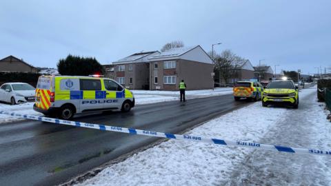 Several police cars can be seen parked along a road in a housing estate. Police tape has been put up to restrict access to the area. Snow is lying on pavements and rooftops and a police officer can be seen standing in the road. 