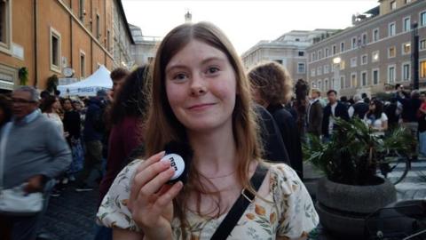 Woman standing in the street holding a small microphone in her right hand and looking in to the camera