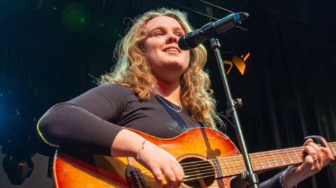A young person, with long blonde hair, holding and playing a guitar, performing into a microphone. It looks like she is on a stage.