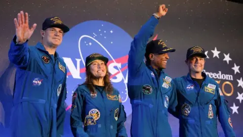 Four astronauts in blue overalls smile and fist pump the air in front of the Nasa logo
