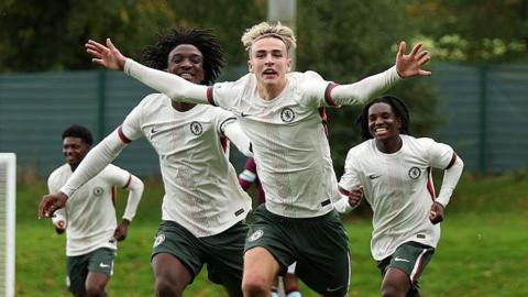 Jesse Derry, wearing Chelsea's light-coloured away kit, has his arms outstretched as he celebrates a goal in an academy game, as his team-mates run to keep up with him