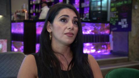 Racha Nassif sitting in the restaurant. She has long dark hair and is wearing earrings and a black top. Behind her a man is standing in the bar area which has purple lighting and drinks displayed on shelves.