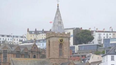 A tradition church, with a clock. The tower on top of the church is made of glass and has a cross. There are houses around the church and a road and cars. 