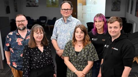 Prof Carol Davenport, left, is wearing a dark blue shirt with patterned images on it. She has dark brown hair. She is smiling and posing for the camera alongside three men and two women. One of the women has pink hair. The man at the back of the photo is wearing a light blue patterned shirt and black glasses. 