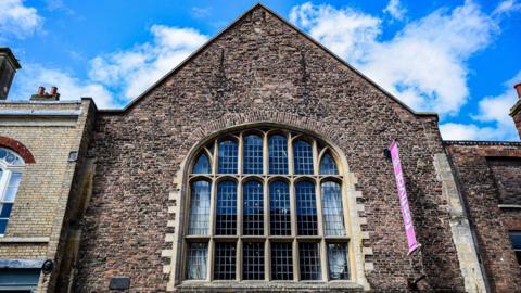 A tall medieval building with a timber façade at the upper end of the structure. There is a blue sky and white clouds above.