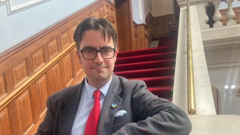 A man with short brown hair and glasses smiling at the camera. He is wearing a jacket with a white shirt and red tie.