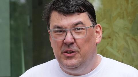 Linehan outside court in May, looking at the camera wearing glasses and a white T shirt.