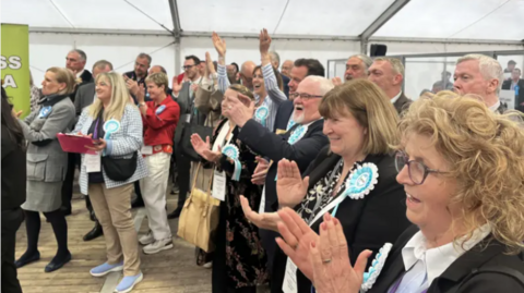 A group of people wearing blue Reform UK rosettes cheer and applaud in a large marquee. 