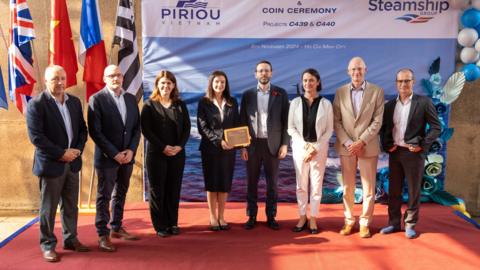 Eight people - five men and three women - line up on a stage in the shipyard in front of a Piriou and Isles of Scilly Steamship Group sign and four flags - including the UK, Vietnam and French flags. A woman stood in the centre is holding a plaque mounted onto wood. 