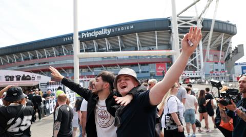 A man in a bucket hat stands with another Oasis fan with their arms in the air outside the Principality Stadium to celebrate the start of the band's tour in Cardiff in June 2025 