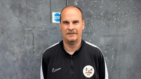 Monty Towers stands, looking at the camera. He has a brown/grey moustache and short brown hair and wears a black and white tracksuit top.