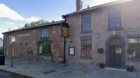 Street view of 'the venue, the sign on the plain brickwork reads, 'The Bootle Arms'. There is an arched doorway set back off the cobbled pavement and a bus stop in front of the building.