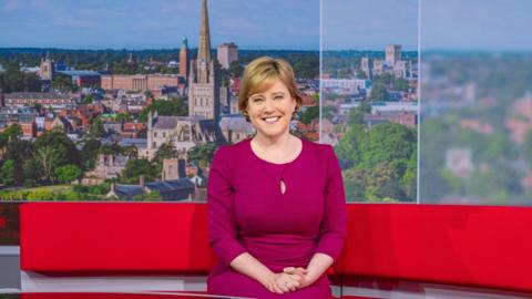 Susie Fowler-Watt, wearing a cerise-coloured dress, sits on the red Look East sofa. She is smiling with her hands clasped. The studio backdrop shows a view of Norwich with landmarks including both cathedrals and City Hall 