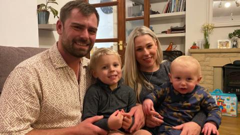 A family of four sitting on a sofa. Dad Charlie is on the left of the photo. He is wearing a cream shirt with a brown pattern. His son Rene is sitting on his knee. He is wearing a black t-shirt. Mum Rebecca is sitting on the right of the sofa, she is wearing a black top and her son Miles is sitting on her knee. He is wearing a top with Christmas reindeers on it.