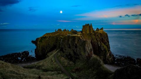 Dunnottar Castle near Stonehaven. The sky is blue and it's getting darker. A ruined castle stands atop a small and tall patch of land surrounded by water with a little path leads back to the mainland, where the photographer is. The moon is out and the sky is streaked with orange.