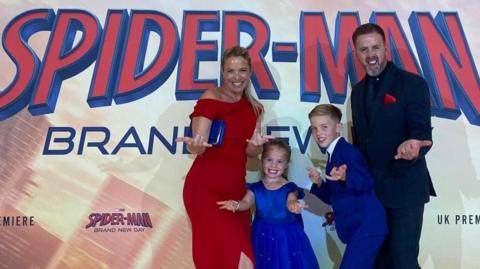 A blond woman in a red dress, man in a black suit, and two children in royal blue do the spider man pose, extending their hands to the camera, in front of the premiere poster.