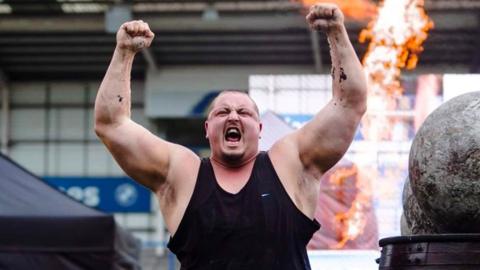 A man with his arms aloft at a strongman competition. He has short hair and is wearing a black vest.