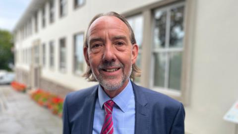 Deputy Steve Falla, a man with grey hair just above his shoulders and a grey beard. He is wearing a light blue shirt, a red tie with white and blue stripes, and a navy blue jacket. In the background is a building with a row of windows and a flower bed in which red flowers are growing.