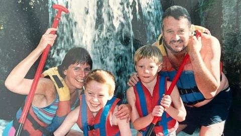 A family of two young boys and two parents stood in front of a waterfall with oars and all wearing red life jackets