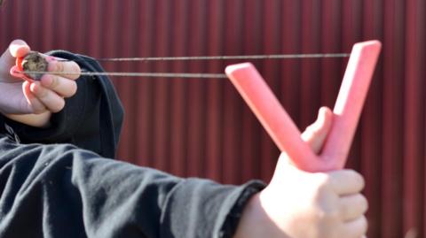 A person, wearing a black jumper, holding a catapult and in position to fire.
