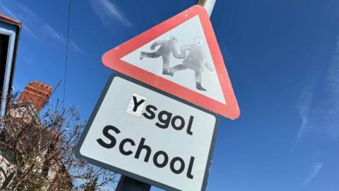 A bilingual school sign in Bangor, Gwynedd. The square sign says "Ysgol" followed by "School" underneath, and above is a red triangle sign that has an image of a child walking, holding the hand of an adult. Behind the sign there are blue skies and a tree with a house behind it to the left.