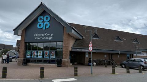 The Co-op in Stornoway is a large building. There is a front entrance with a glass frontage and peaked roof. The word "Co-op" is in blue letters and below it words in Gaelic to say it is the Co-op in Stornoway. Behind the entrance is a long building with a peaked roof.
