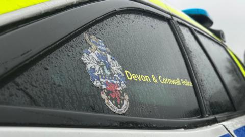 A close up picture of a Devon and Cornwall Police car. The forces logo is visible on the window of the car. There is lot of rain water on the window. 